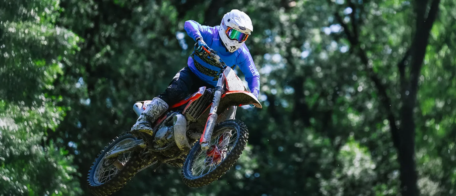 Een motorrijder in een blauwe, lange mouwen racepak en witte helm springt op een motorfiets door een bos.