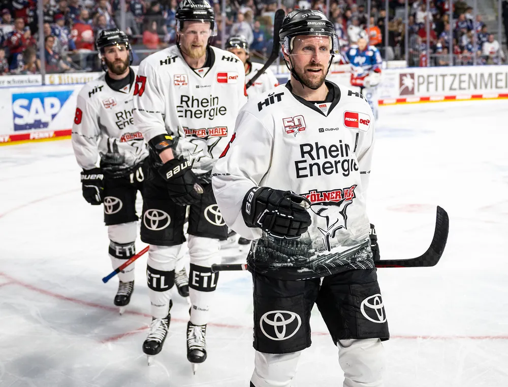 Drie ijshockeyspelers in witte jerseys met zwarte logo's en grafische ontwerpen staan op het ijs.