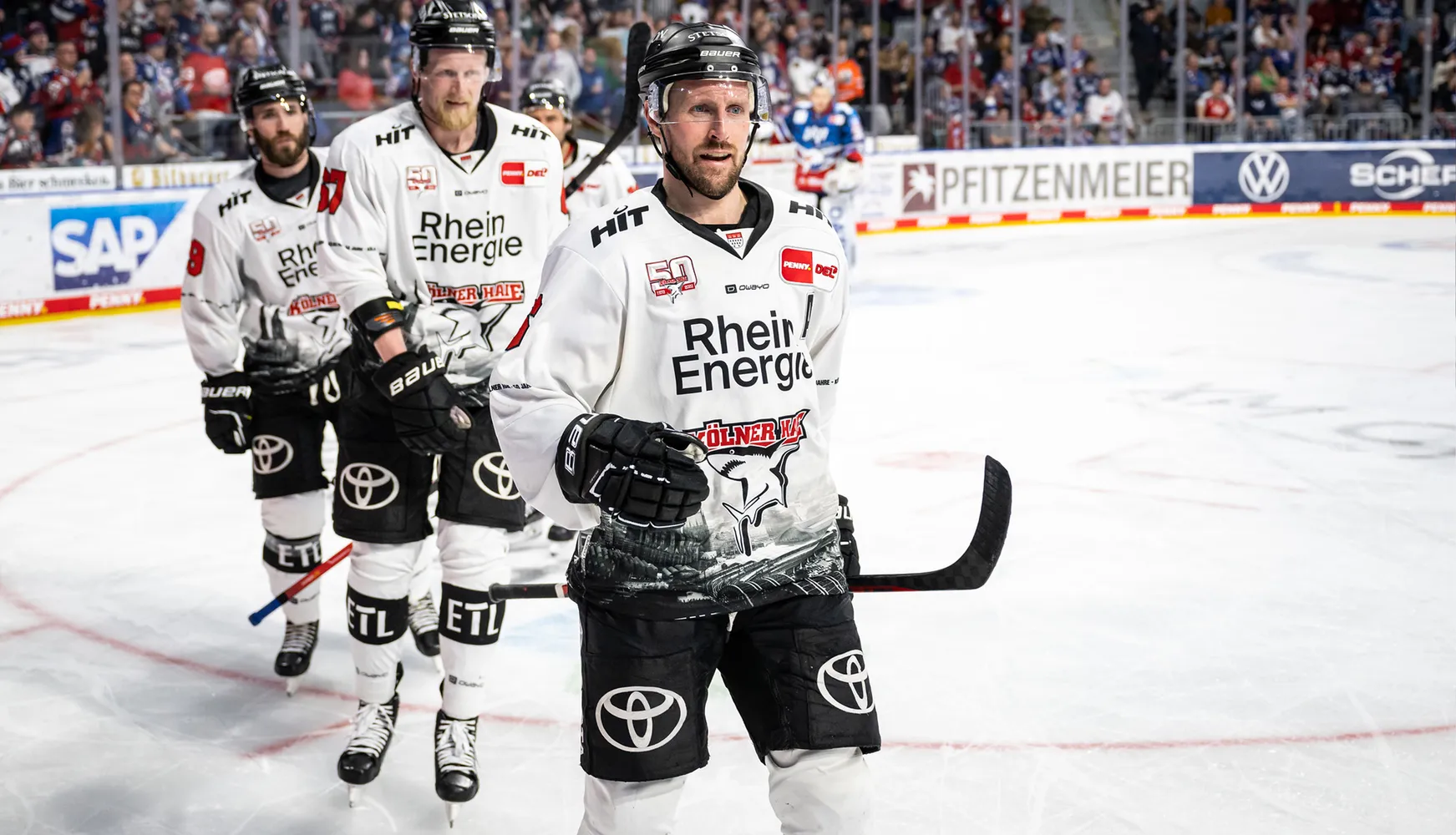 Drie ijshockeyspelers in witte shirts met verschillende nummers staan op het ijs tijdens een wedstrijd.