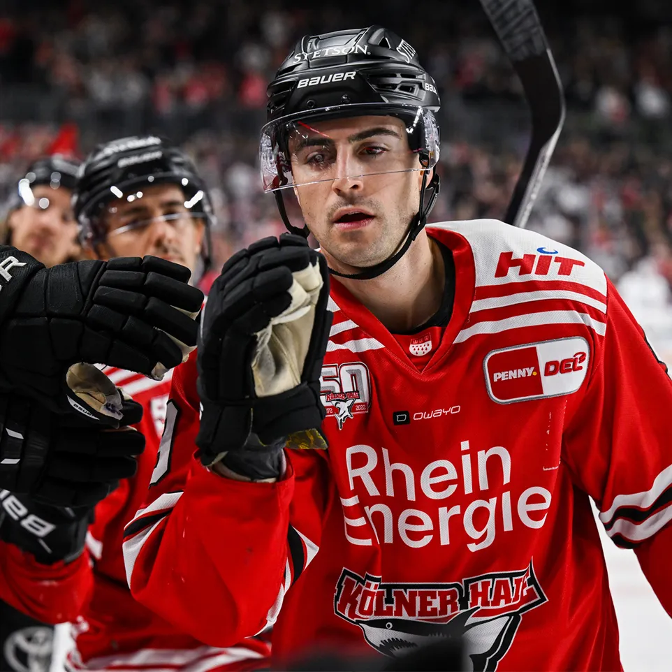 Een hockeyspeler in een rood shirt met witte strepen en logo's, die op het ijs staat en zijn handschoenen opheft.