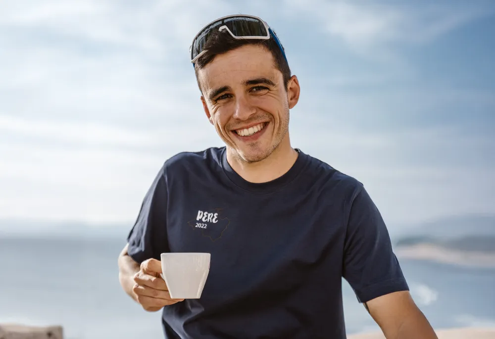 Een lachende man in een blauw T-shirt houdt een kopje vast, met een prachtig kustlandschap op de achtergrond.