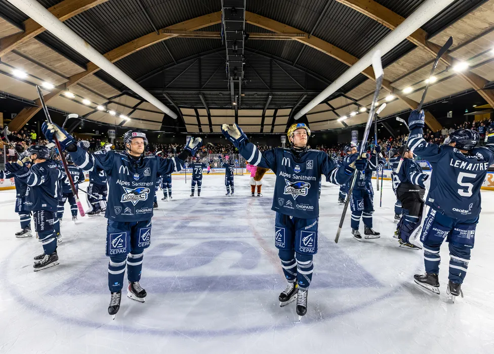 IJshockeyspelers in blauwe shirts staan op het ijs en vieren met opgestoken sticks voor een juichende menigte.