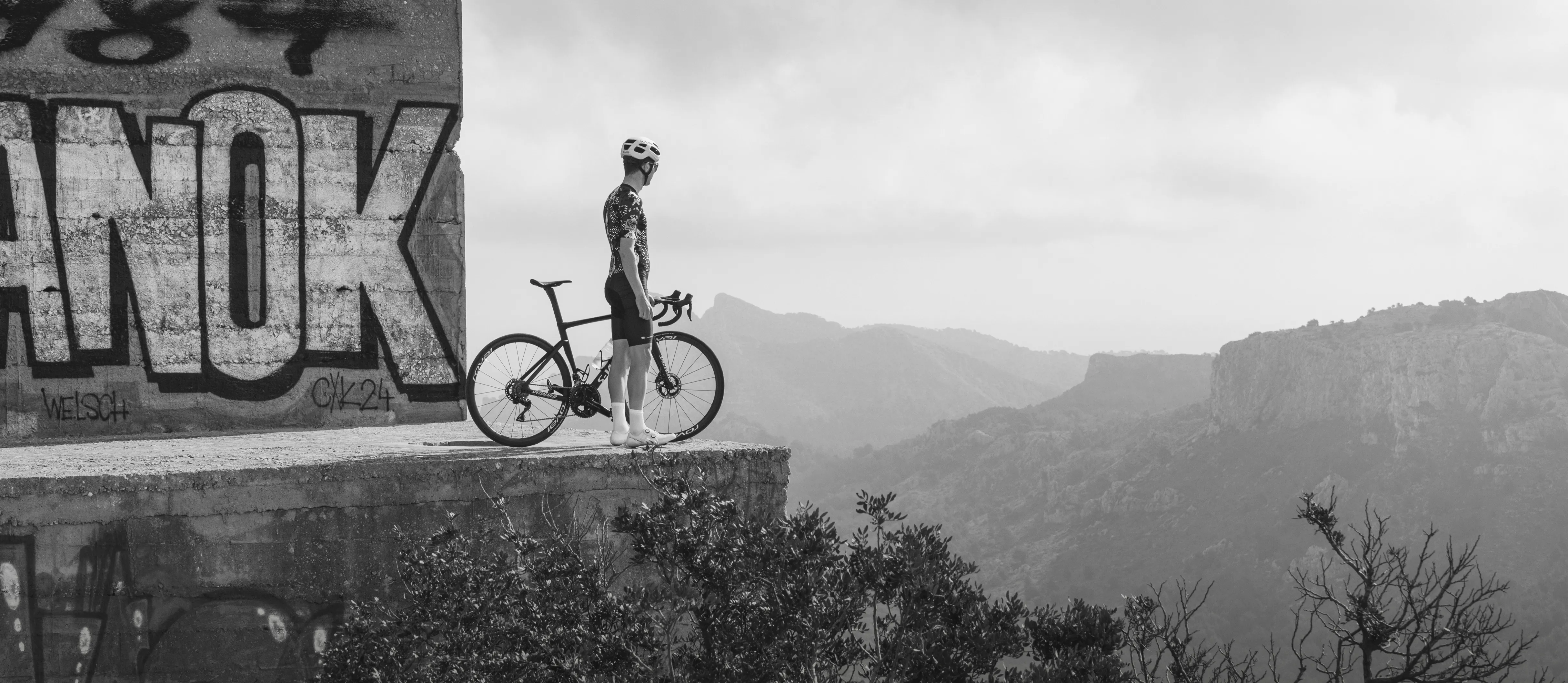 Een fietser in een gemusterde sportkleding staat met zijn racefiets op een betonnen platform naast een muur met graffiti en kijkt uit over een bergachtig landschap.