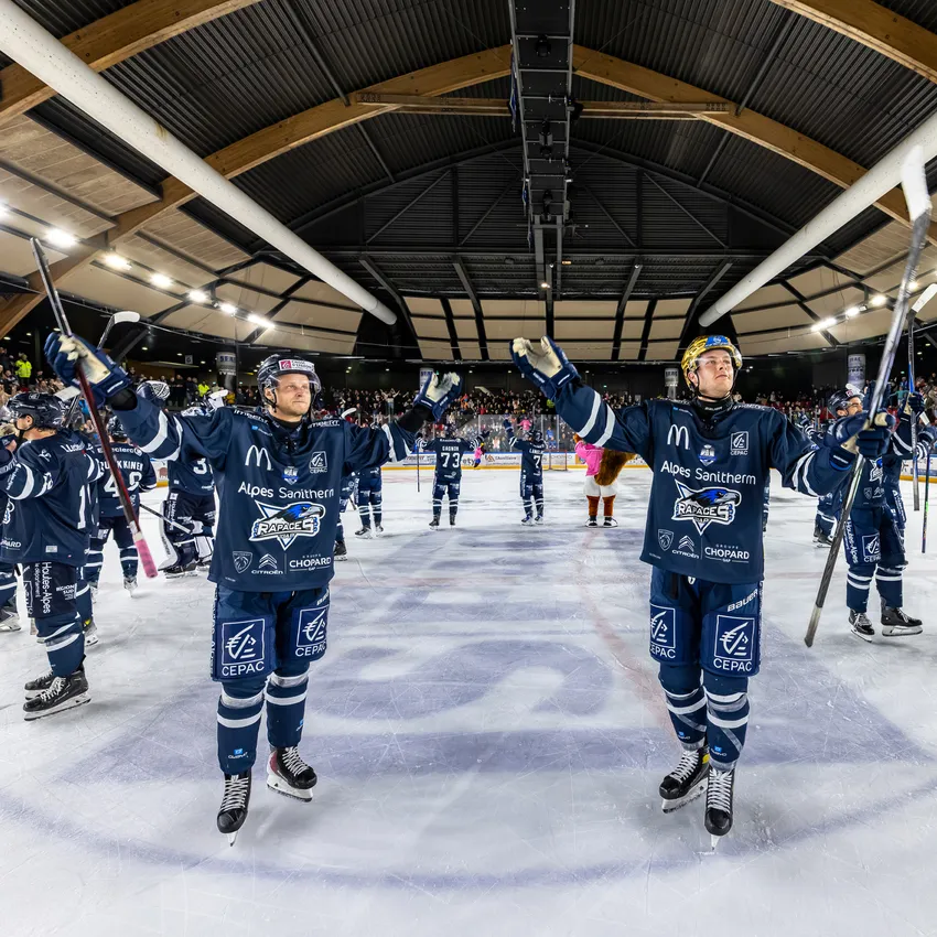 Twee ijshockeyspelers op het ijs die vieren met hun sticks, terwijl andere spelers en toeschouwers op de achtergrond zichtbaar zijn.