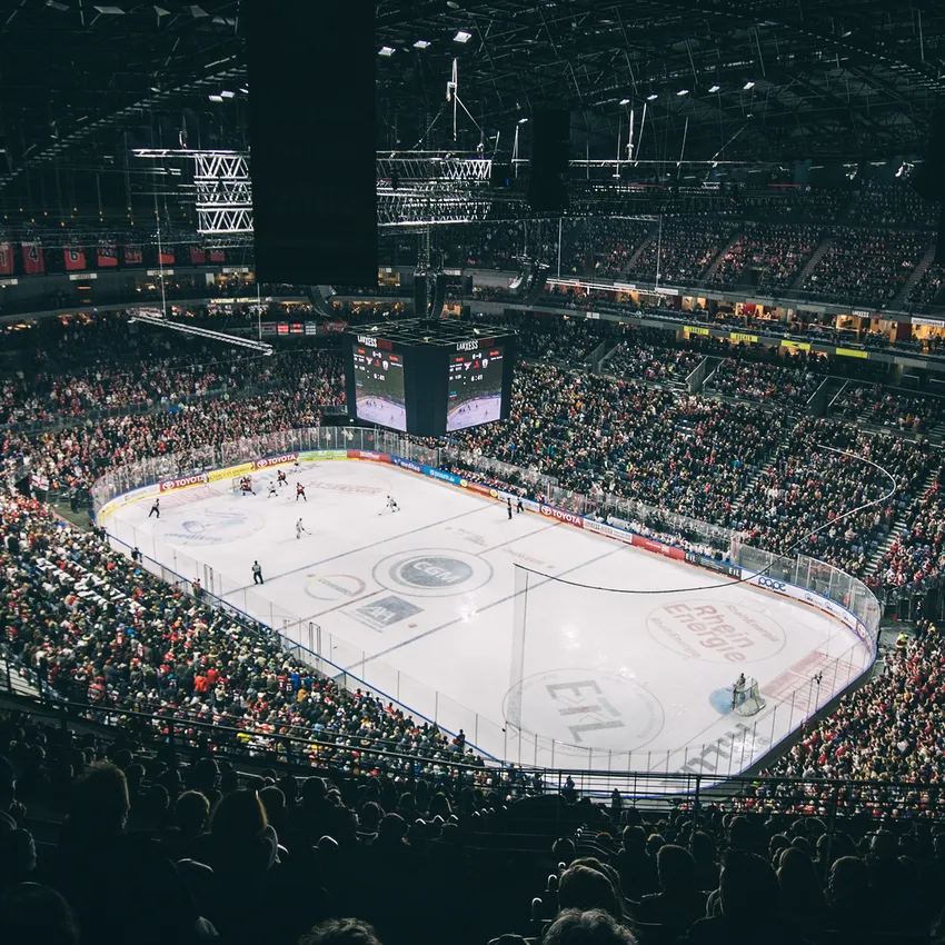 Een volle ijshockeyarena tijdens een wedstrijd, met spelers op het ijs en een groot scorebord op de achtergrond.