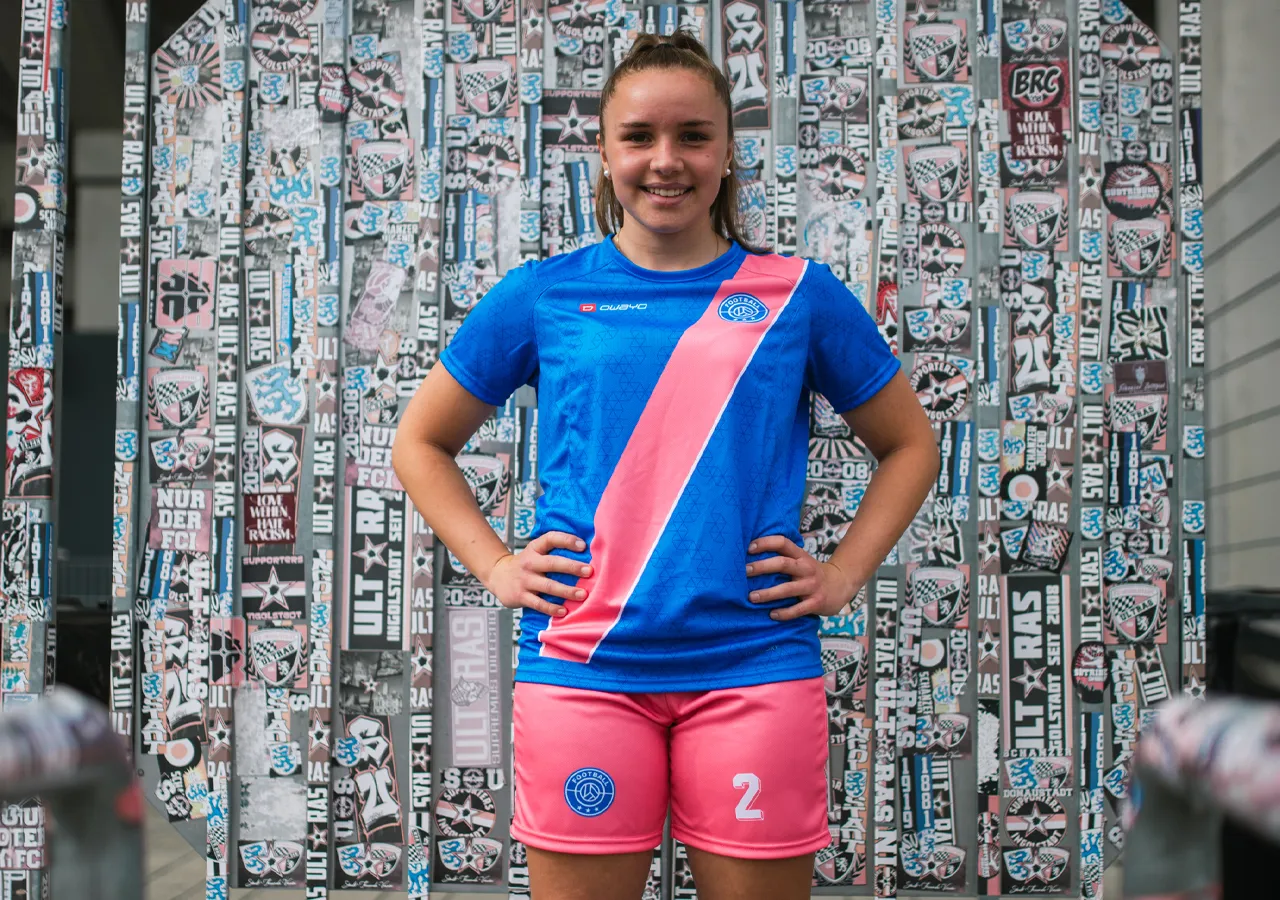 Een jonge vrouw draagt een blauw voetbalshirt met een roze diagonale streep en een roze short met het nummer 2, voor een muur bedekt met grafieken en stickers.