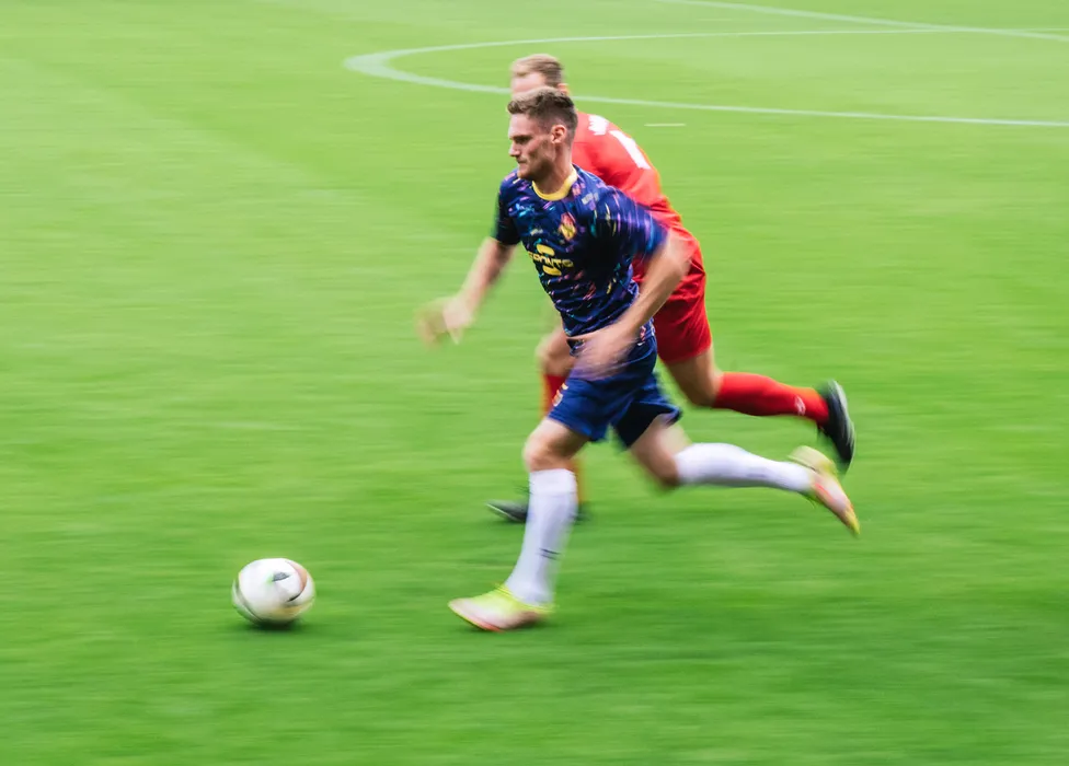 Een voetballer in een blauw shirt met kleurrijke patronen rent snel over het groene veld terwijl een andere speler dicht achter hem aanloopt.