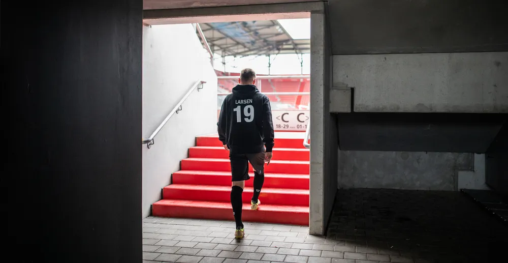 Een atleet in een zwarte hoodie met het nummer 19 en zwarte shorts die een rode trap in een stadion oploopt.