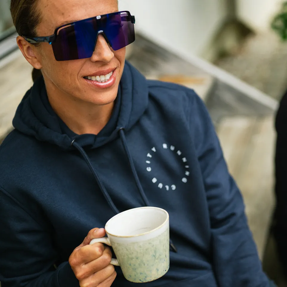 Een persoon in een donkerblauwe hoodie met een logo glimlacht naar de camera terwijl hij een mok vasthoudt.