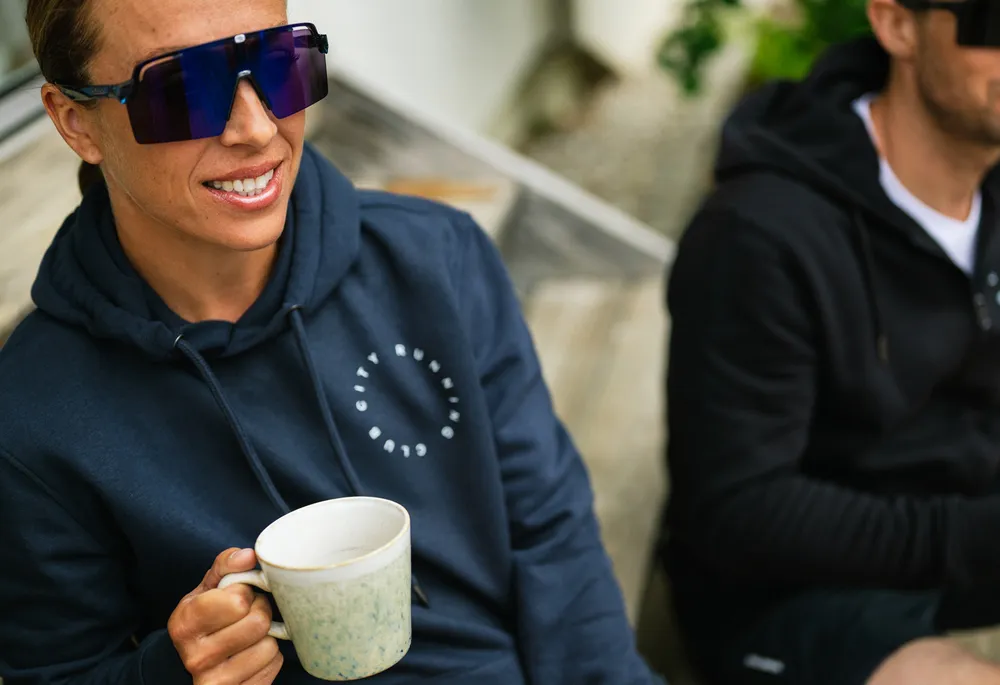 Een persoon in een blauwe hoodie met een logo houdt een kop vast terwijl hij op een houten bank zit.