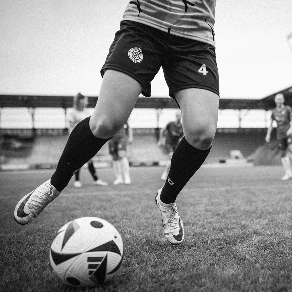 Een speelster in korte sportbroeken en kousen die een voetbal dribbelt op een grasveld.