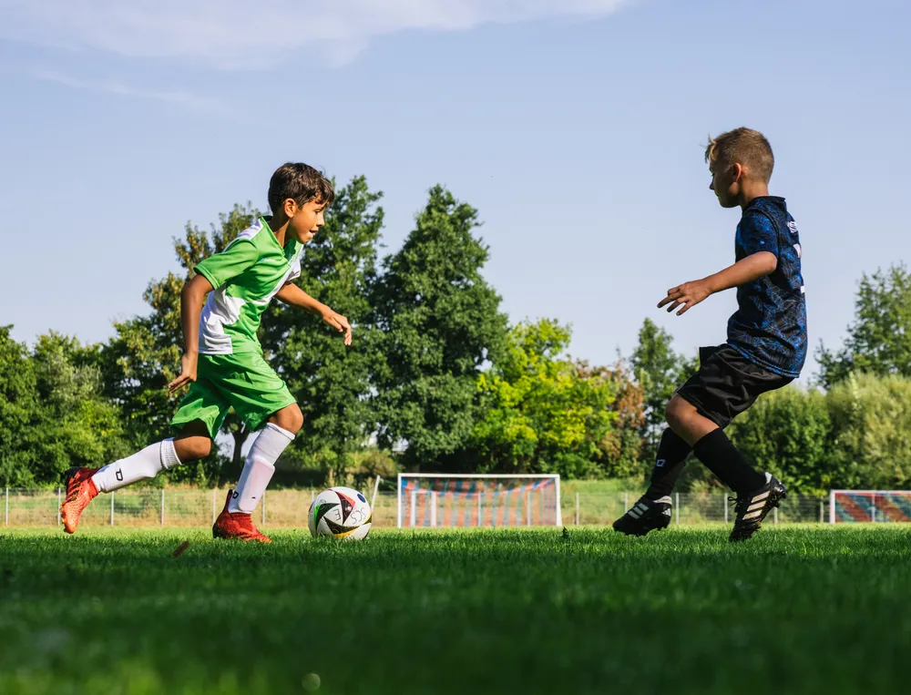 Twee kinderen spelen voetbal op een groen veld, de een in een groen shirt en de ander in zwart, terwijl ze de bal achternazitten.