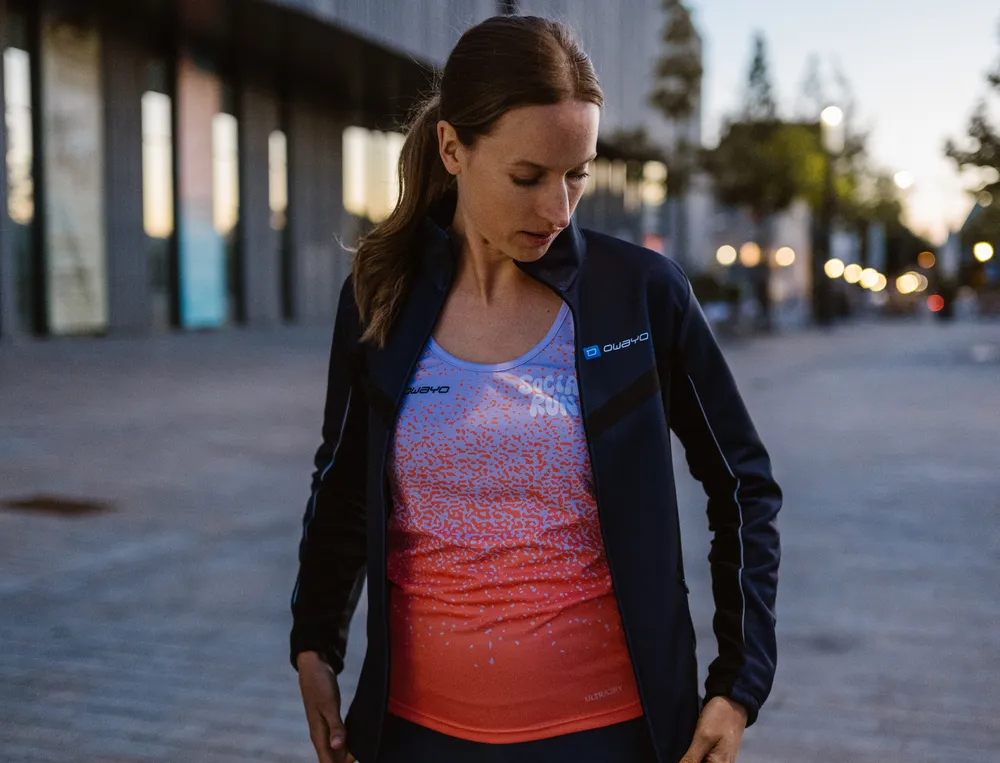 Een vrouw in sportkleding, bestaande uit een lichte jas en een kleurrijke top, staat in een stedelijke omgeving en kijkt nadenkend naar haar outfit.