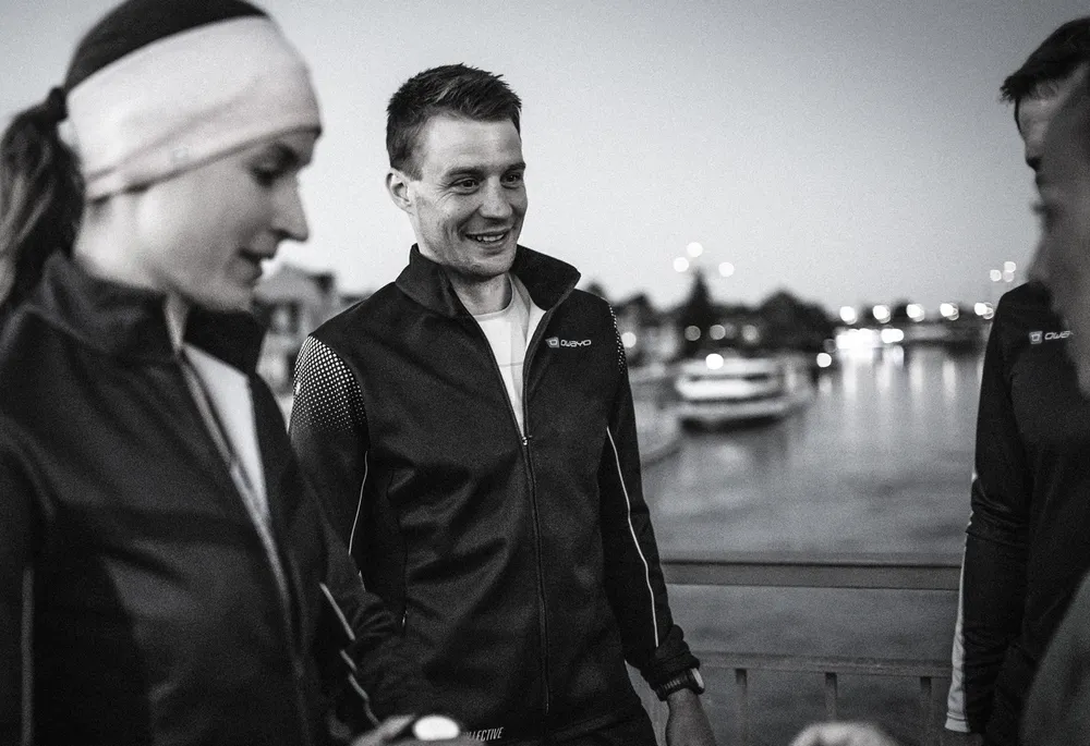 Een groep atleten in donkere sportjassen staat aan de waterkant tijdens de schemering.