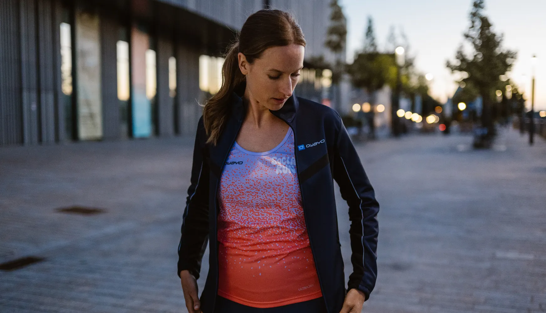 Een vrouw in een sportieve outfit met een zwarte jas en een kleurrijke, grafische tanktop, staande op een stoep.