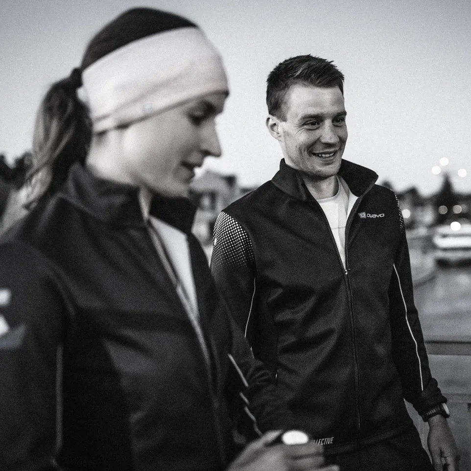Twee atleten in moderne zwarte jassen staan buiten, glimlachend en klaar voor training.
