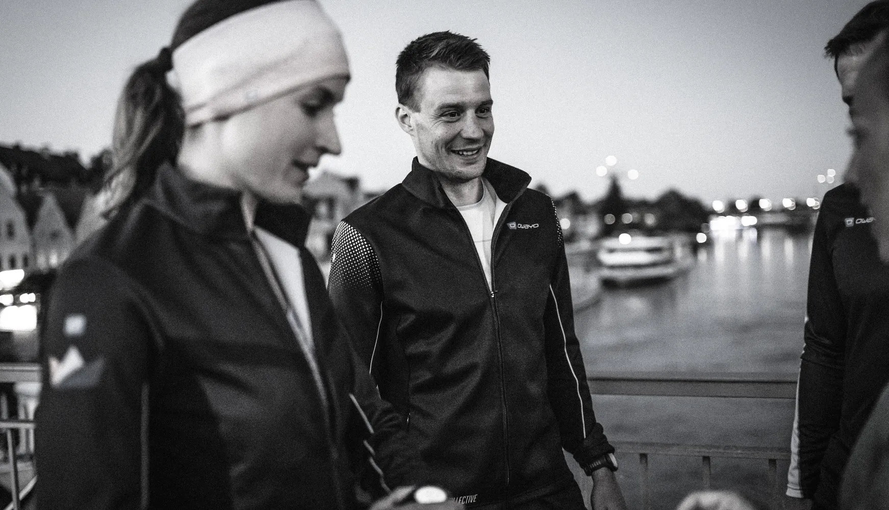 Twee mensen in sportieve jassen staan aan de waterkant, de een met een hoofdband die nadenkend kijkt, terwijl de ander glimlacht.