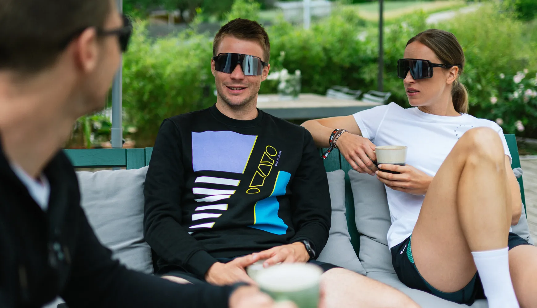 Drie personen zitten ontspannen op een bank, waarvan er één een zwart sweatshirt met een kleurrijk ontwerp draagt en de ander een wit t-shirt, terwijl ze drankjes vasthouden.