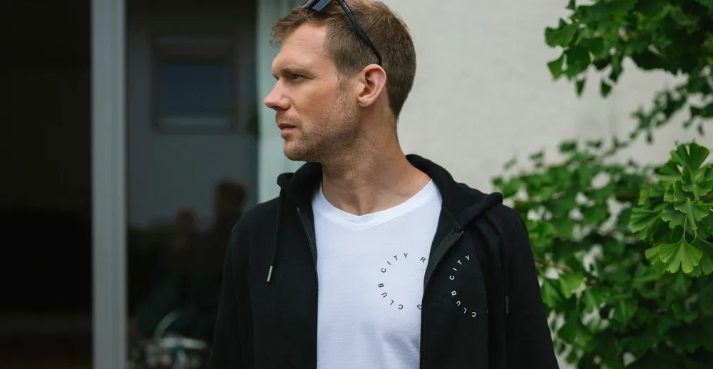 Een man staat zijwaarts en draagt een zwarte hoodie over een wit T-shirt met een print, omringd door planten.