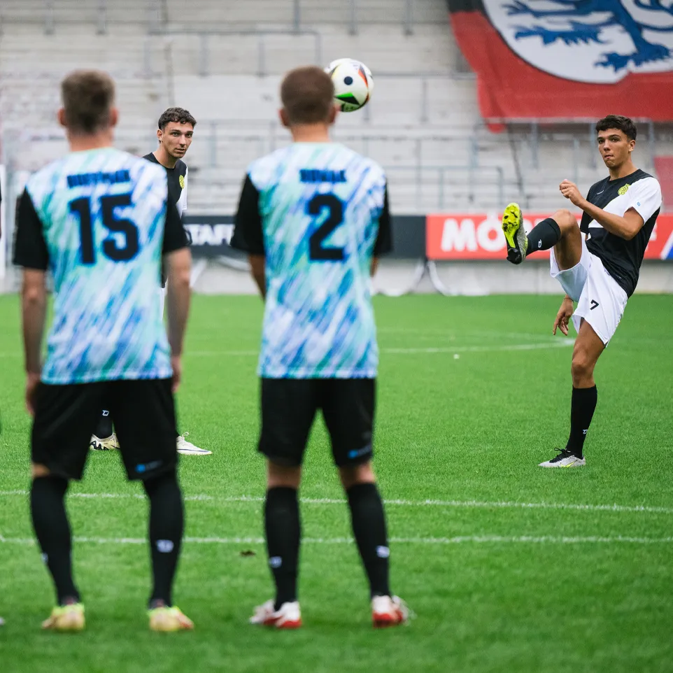Drie voetballers in kleurrijke shirts staan op het veld en kijken naar een andere speler die met een bal jongleert.