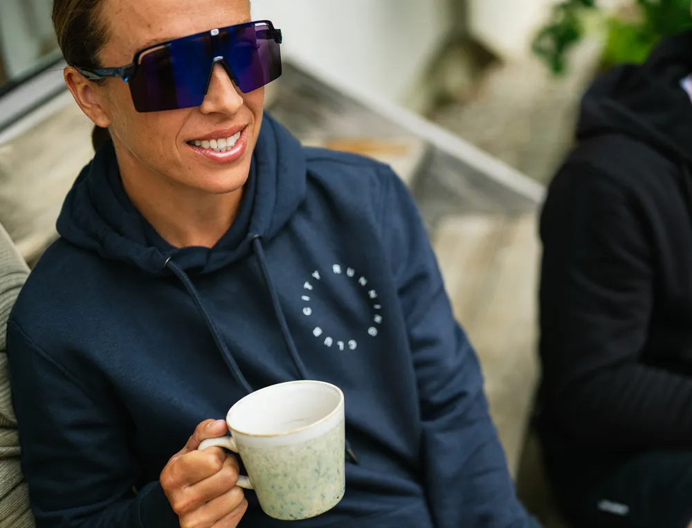 Een persoon in een marineblauwe hoodie met een logo houdt een kop koffie vast en draagt grote zonnebrillen.