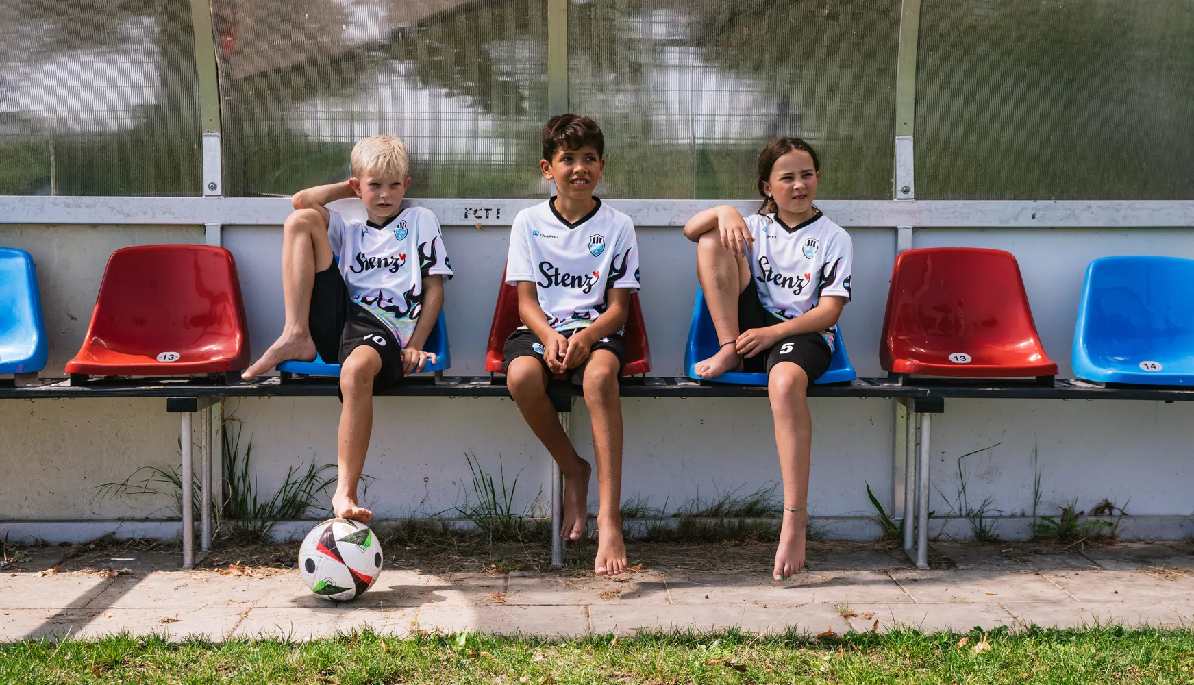 Drie kinderen in gepersonaliseerde voetbalshirts zitten op een bank met een voetbal op de grond.