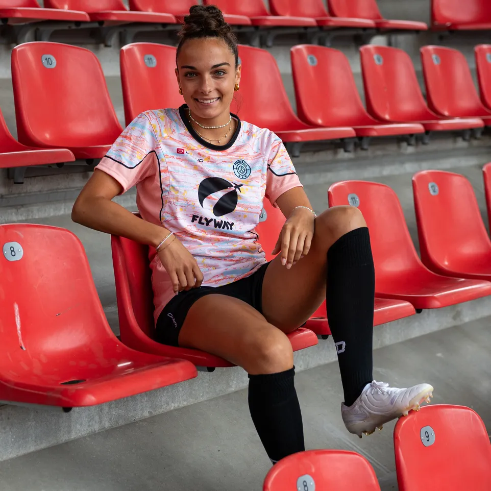 Een glimlachende vrouw zit in een stadion op een rode stoel, gekleed in een kleurrijk voetbalshirt en zwarte sokken.