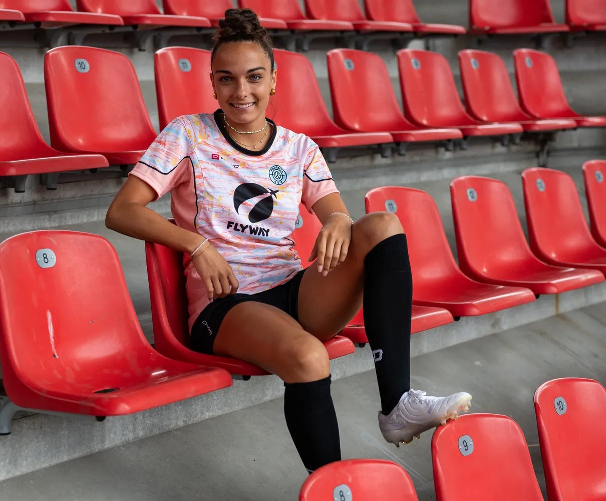 Een jonge vrouw zit op een rode stadionstoel in een kleurrijk voetbalshirt en zwarte sokken.