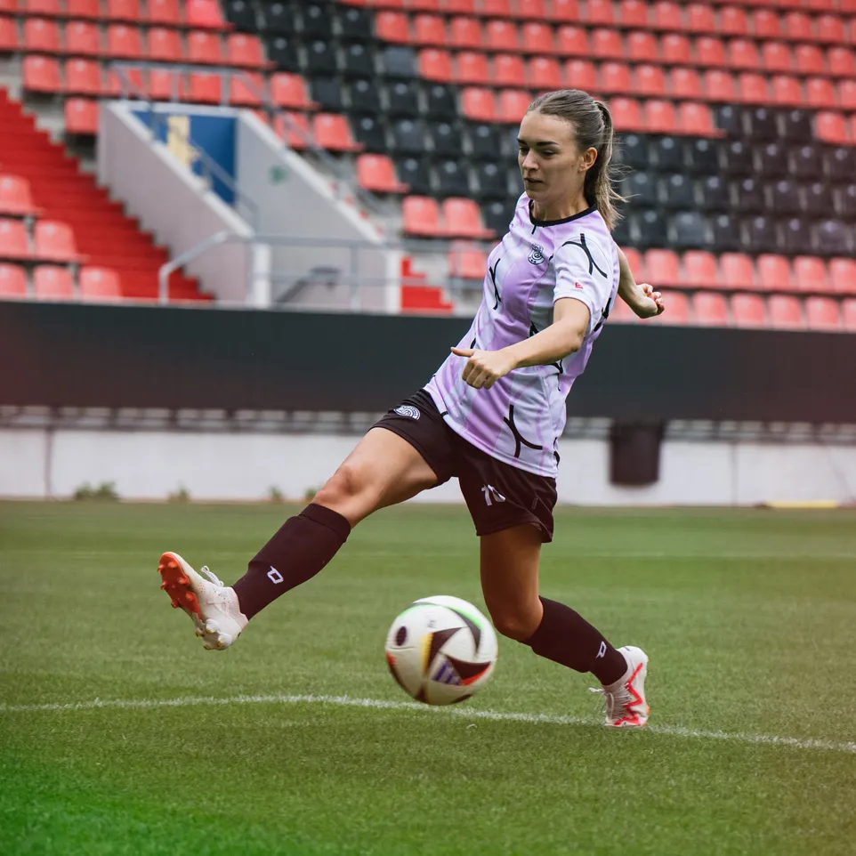 Een vrouwelijke speler schopt een voetbal op een stadionveld, gekleed in een paarse trui en zwarte shorts.