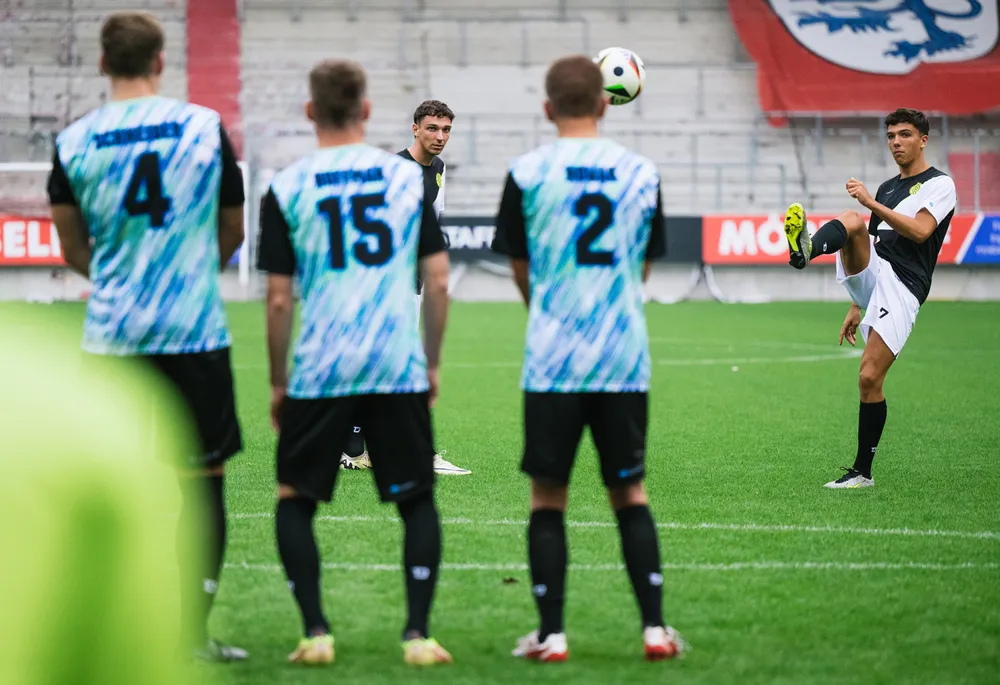 Een groep voetballers in kleurrijke shirts staat op het veld terwijl een speler zich voorbereidt om een vrije trap te nemen.