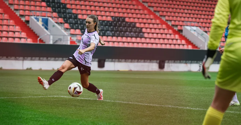 Een vrouwelijke speler in een paars-wit voetbalshirt schopt de bal op een stadionveld, met een doelman in gele uitrusting op de achtergrond.