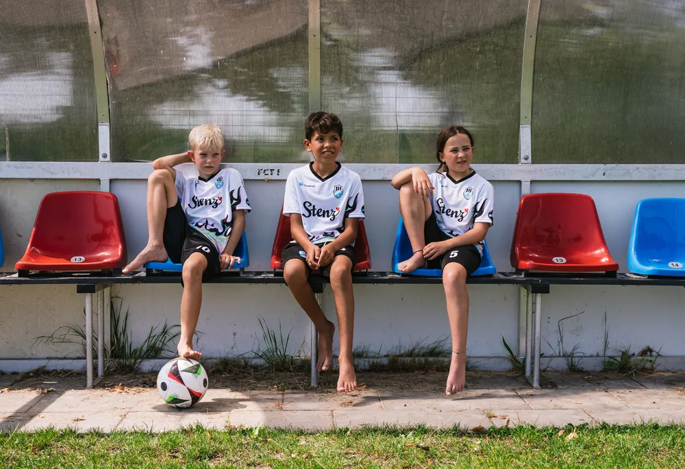 Drie kinderen in sportieve voetbalshirts zitten op een bank naast een voetbal.