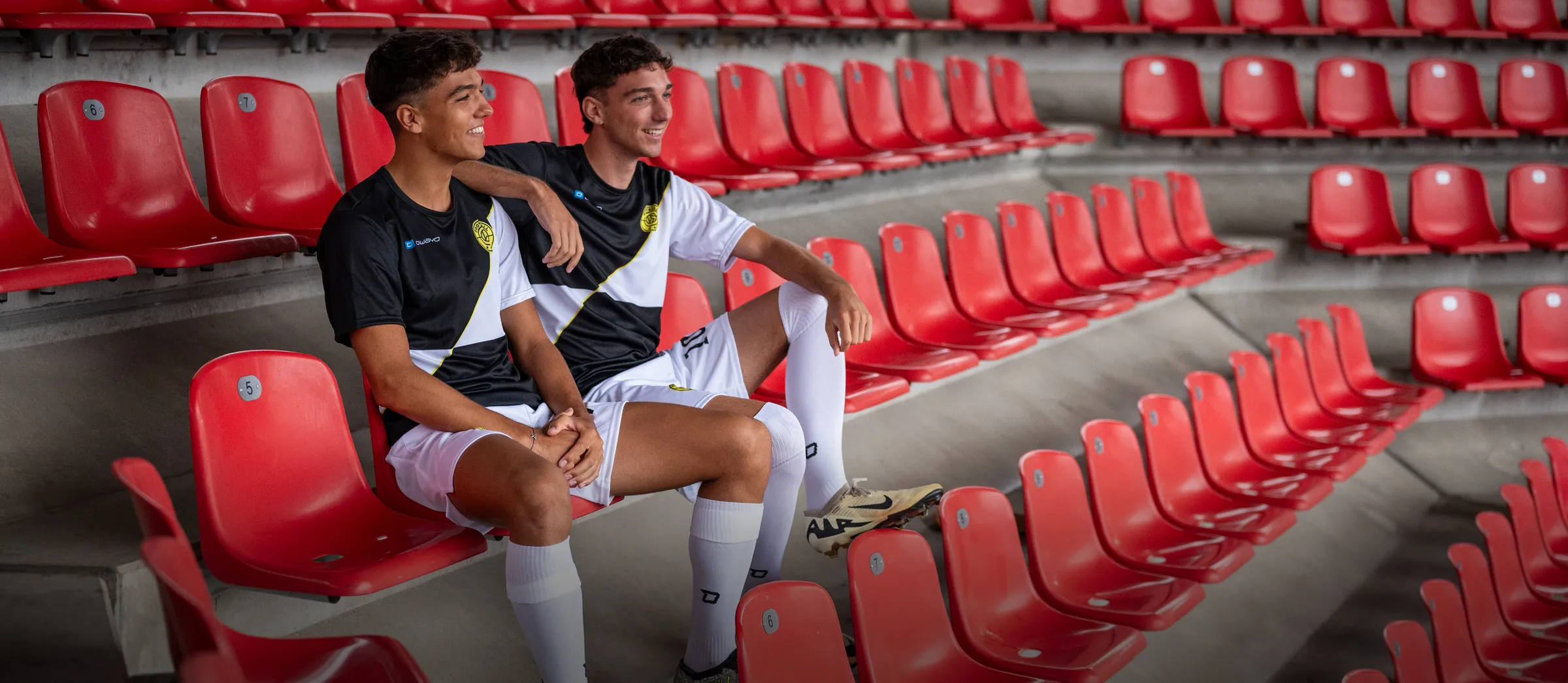 Twee jonge mannen in sportieve voetbaloutfits zitten casual op rode stadionstoelen.