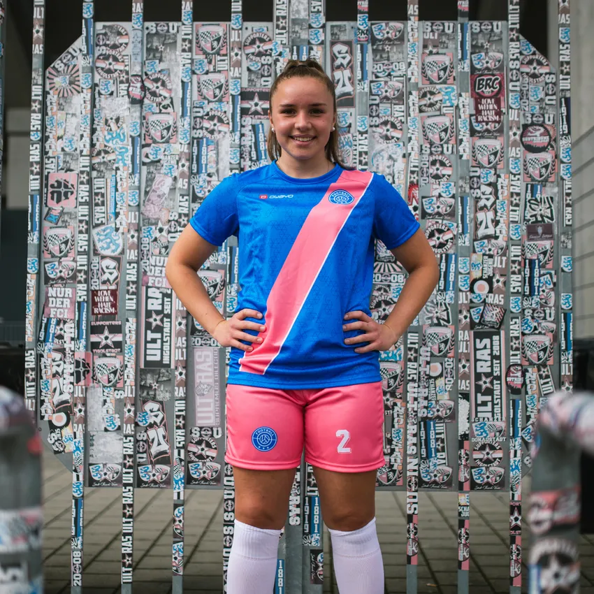 Een jong meisje in een blauw voetbalshirt met een roze streep en roze shorts staat voor een muur bedekt met stickers.