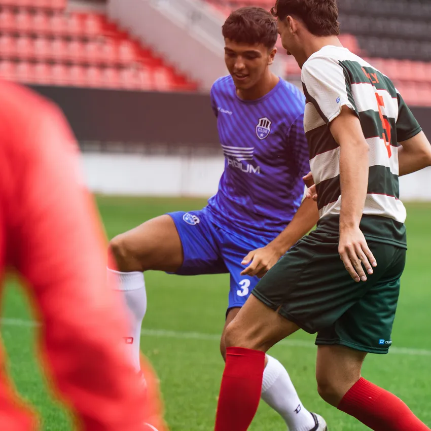 Twee voetballers in actie, de een in een blauw shirt en de ander in een gestreept groen en wit shirt, staande op een voetbalveld.
