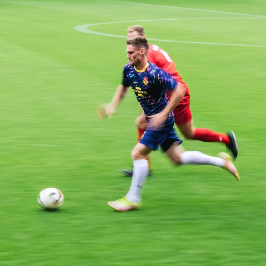 Een voetballer in een kleurrijk shirt rent over het veld terwijl een andere speler hem achterna zit.