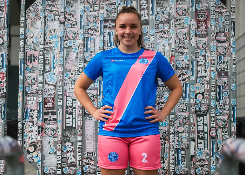 Een jonge sporter poseert in een blauw voetbalt-shirt met een roze streep en roze shorts voor een kleurrijke muur bedekt met stickers.