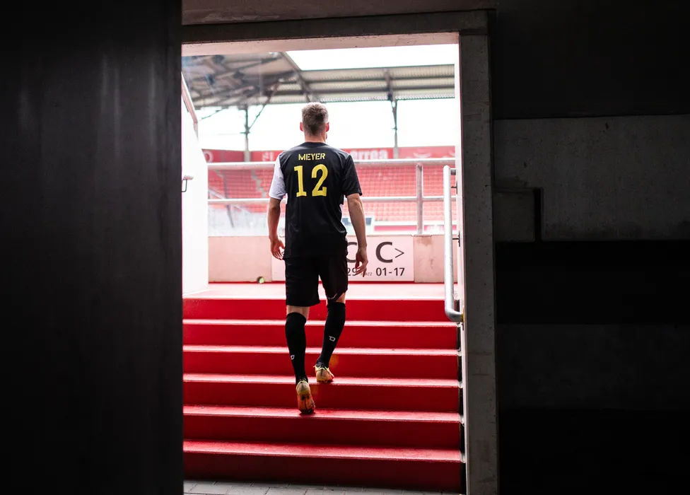 Een voetballer in een zwart shirt met het nummer 12 loopt de rode trappen in het stadion op.