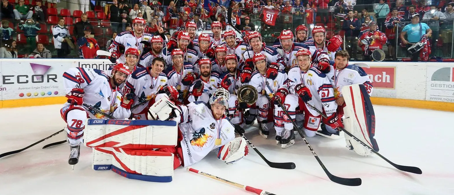 Een ijshockeyploeg in witte shirts met rode en blauwe accenten poseert op het ijs, omgeven door fans, terwijl de spelers medailles dragen en een beker vasthouden.