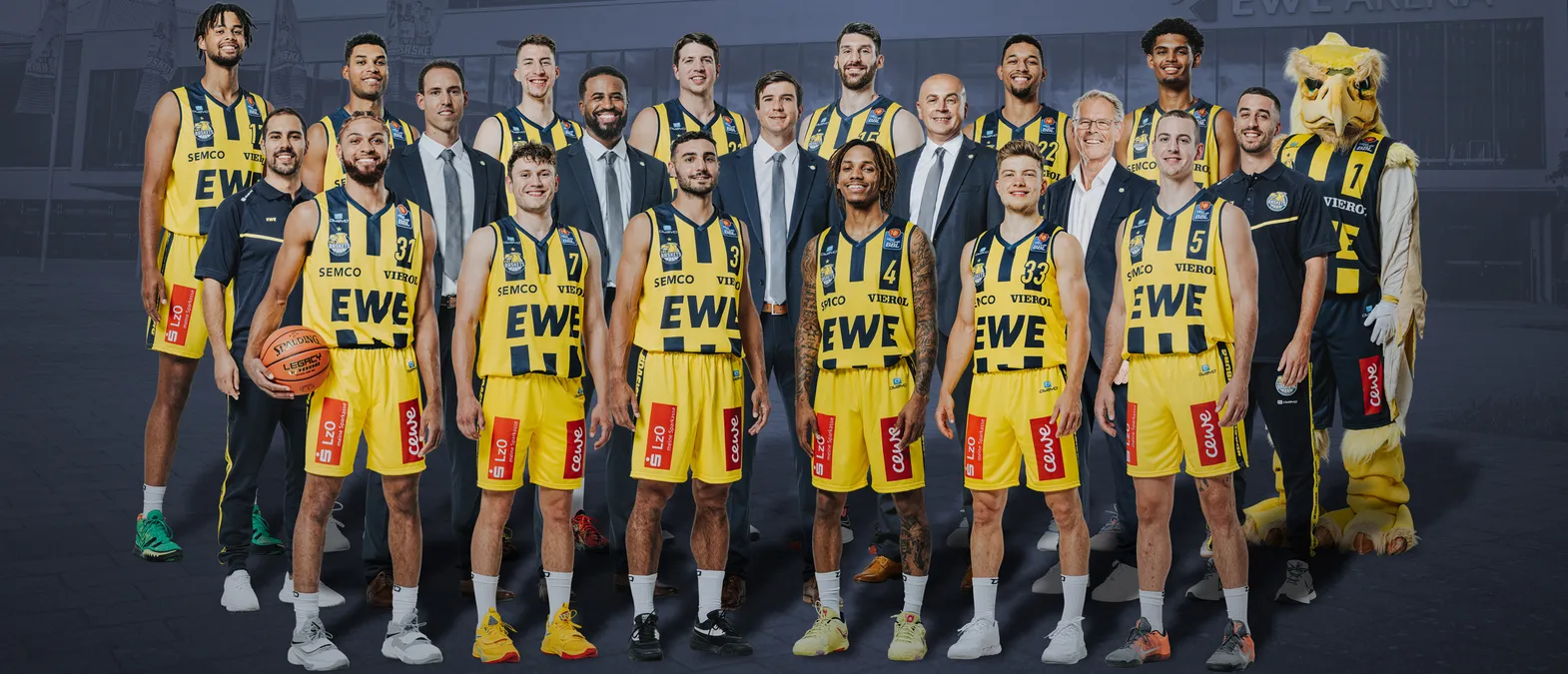 Een groepsfoto van een basketbalteam in geel-zwarte shirts met de sponsors 'EWE' en 'SEMC', een mascotte en een basketbal, voor een gebouw.