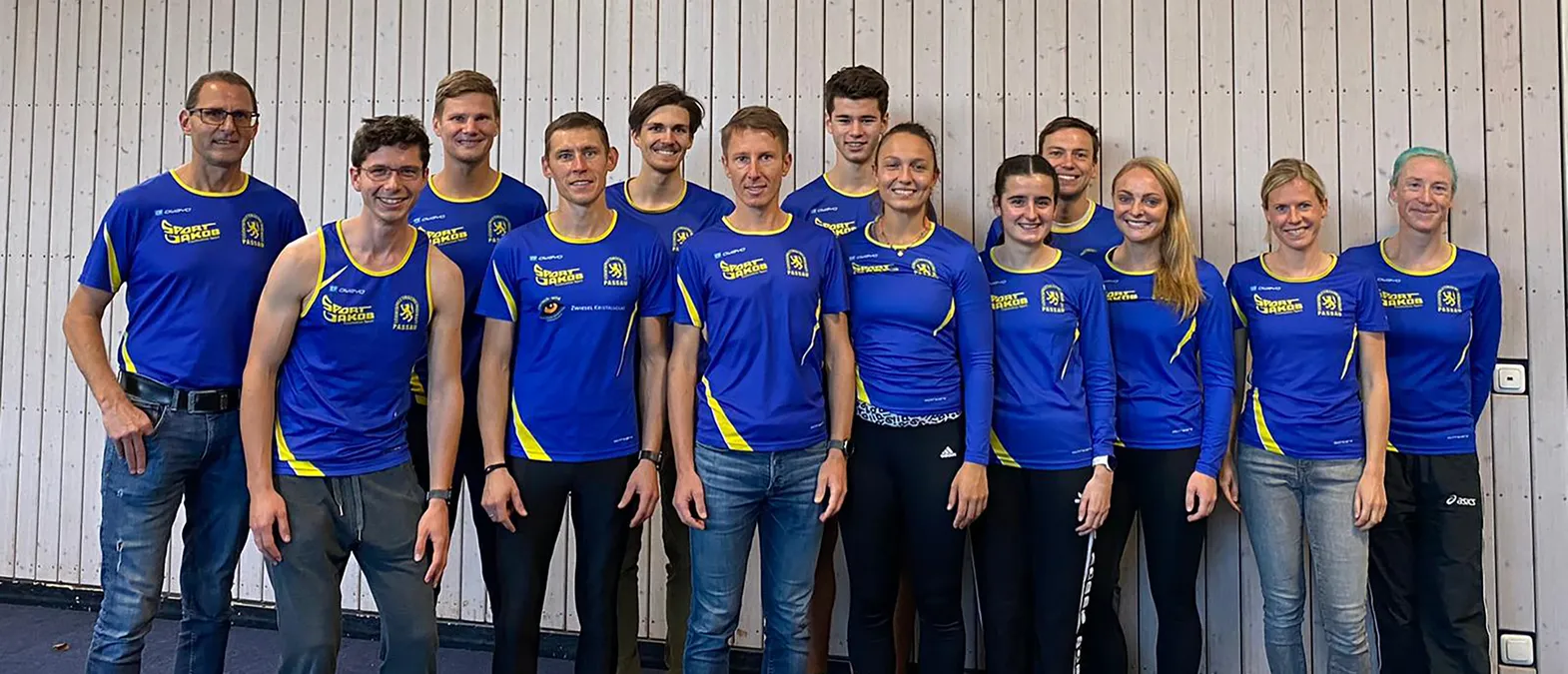 Een groep mannen en vrouwen in blauwe en gele sportkleding met het logo 'Sport Jakob Passau' poseert voor een licht houten wand.