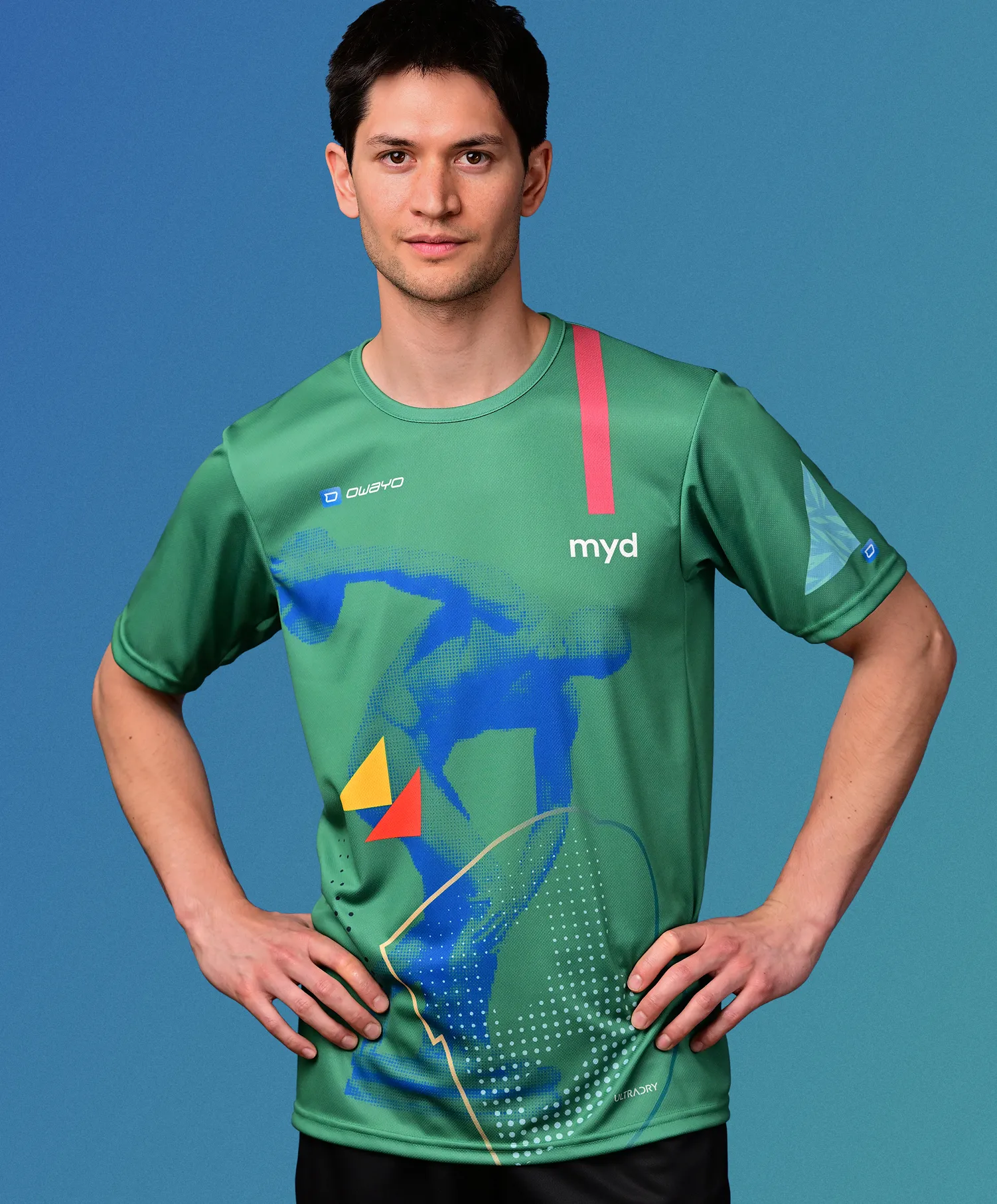 Een man in een groen aangepast sportshirt met een blauw, geel en rood geometrisch ontwerp dat een sportfiguur voorstelt, staat voor een blauwe achtergrond.