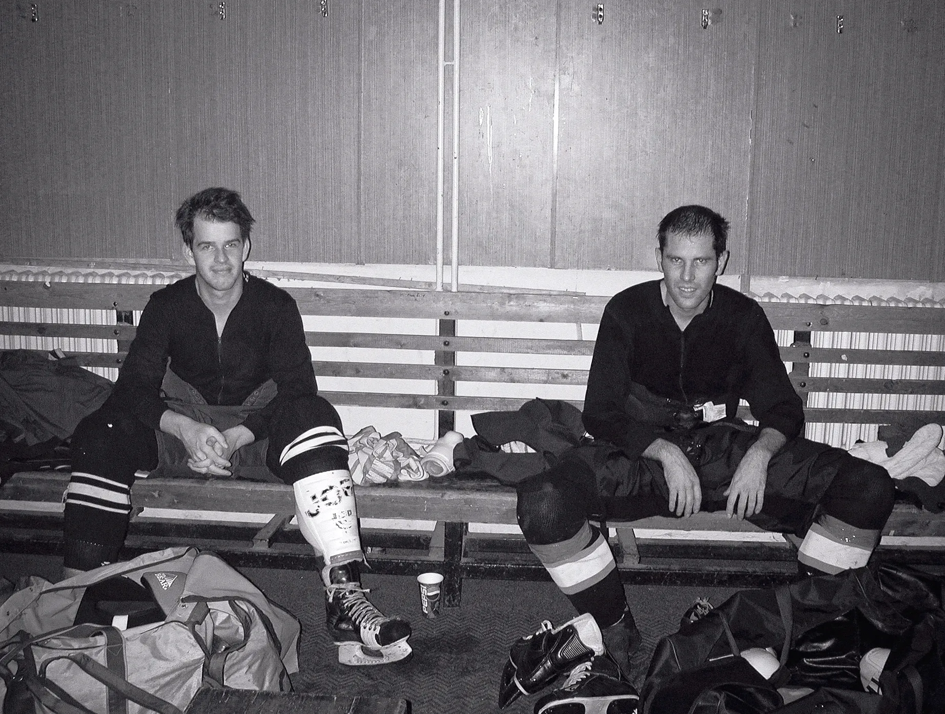 Twee ijshockeyspelers in volledige uitrusting zitten op een houten bank in een kleedkamer, omringd door sporttassen en schaatsen.
