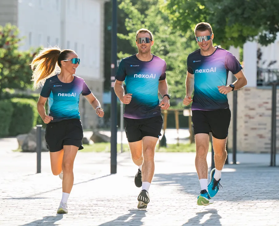 Drie atleten, twee mannen en een vrouw, joggen in bijpassende kleurrijke hardloopshirts en shorts op een geplaveide weg.