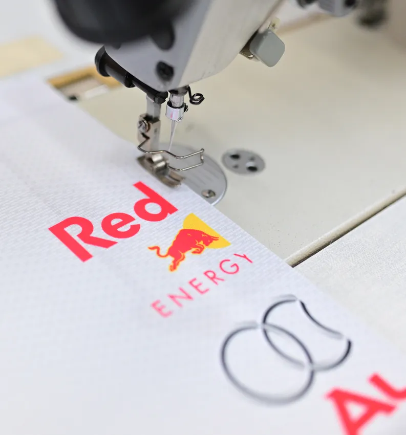 Een naaimachine naait een wit sportkledingstuk met het rode Red Bull-logo.