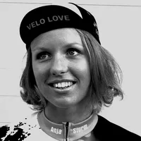 Een vrouw met kort blond haar draagt een zwarte fietshelm met de tekst 'VELO LOVE' en een aangepast fietspak met de naam 'Anja Sturm'.