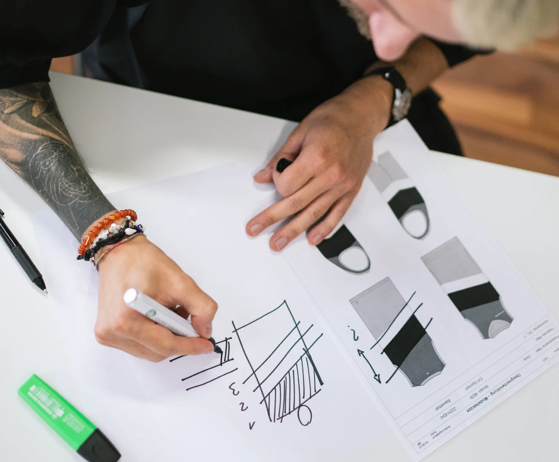 Een persoon met een tatoeage op de arm tekent met een marker een ontwerp op een vel papier, terwijl technische tekeningen van sportkleding op tafel liggen.