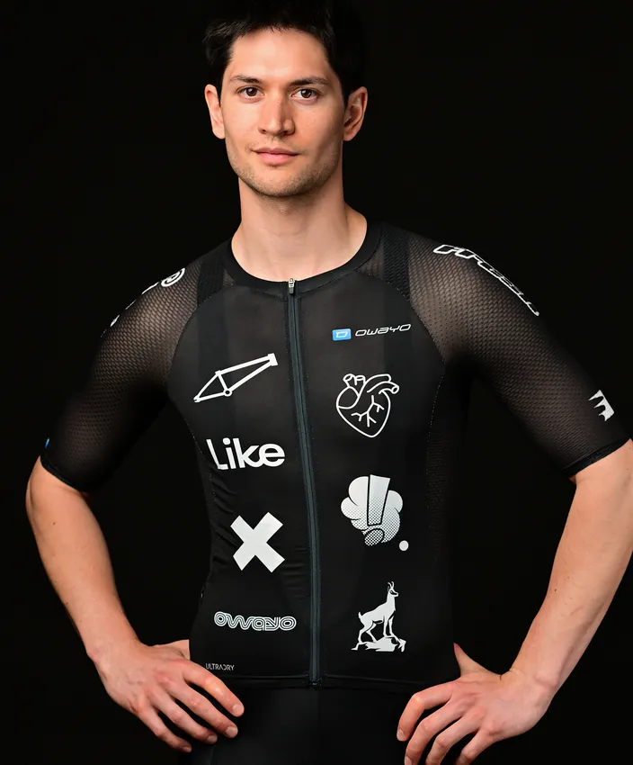 Een jonge man draagt een nauwsluitend zwart sportshirt met verschillende witte logo's en ontwerpen, poserend tegen een zwarte achtergrond.