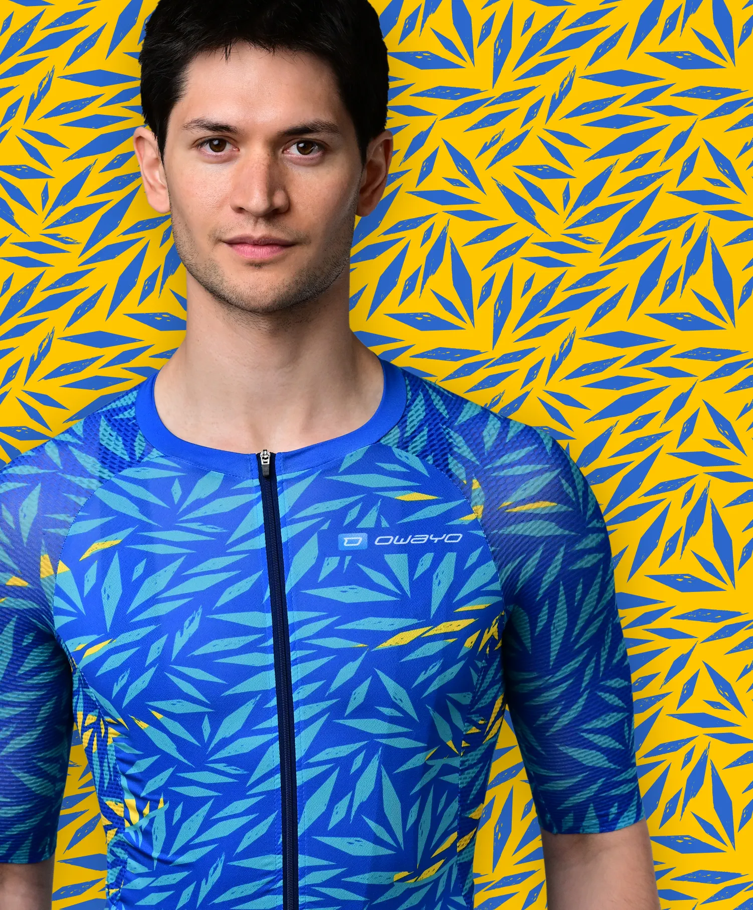 Een man draagt een blauw, korte mouwen sportshirt met een blauw en geel bladpatroon dat doet denken aan een tropische plant, tegen een gele achtergrond met blauwe bladpatronen.
