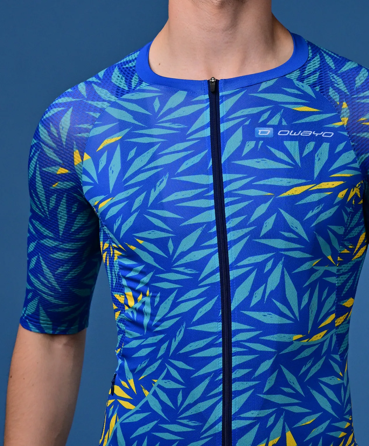 Een man draagt een blauw, korte mouwen sportshirt met een tropisch bladpatroon in turkoois en geel en een rits aan de voorkant.
